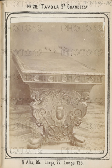 Table with decorated legs, No 29 Tavola, photograph, page, inscription: '2A. Grandezza, N Alta, 85. Larga, 77. Lunga, 125.', inscription: 'Ies. 70', c. 1878 - c. 1885, photographer, paper, albumen print, height 126 mm x width 96 mm, Photo on unnumbered page., table, A sepia-toned photograph of an ornately carved table top and its pedestal, showing a flat rectangular surface with a decorative edge and a frieze with circular motifs, supported by a richly carved central leg featuring symmetrical scrollwork, a shield-shaped cartouche, floral elements, and two carved female heads with wings, resting on claw-like feet and set on a patterned floor, all framed by a printed border and page margins with printed headings and marginal markings.