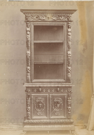 Kast, No 7 Mobile, photograph, page, inscription: 'Alto, 218. Largo, 92. Fondo, 50.', inscription: 'Ia. 130', c. 1878 - c. 1885, photographer, paper, albumen print, height 135 mm x width 96 mm, Photo on unnumbered page, cupboard, A tall, ornate wooden cabinet with carved decorative molding and relief panels is shown against a plain backdrop, the upper section featuring an open display area with shelves framed by carved borders and fluted pilasters adorned with sculpted faces and floral motifs, the lower section composed of paneled doors with raised, circular medallion carvings and intricate scrollwork, the base resting on carved feet and embellished with repeated ornamental patterns, the surface showing a uniform, aged tone and slight surface blemishes.