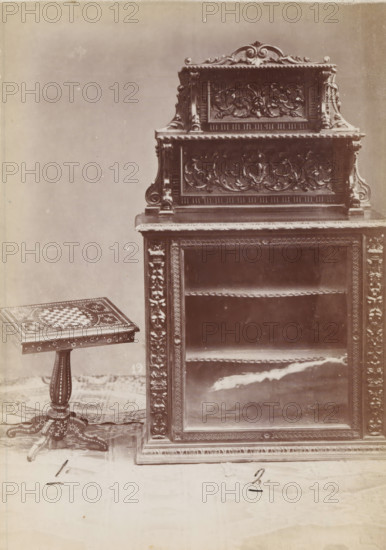 No 12 Etager, Wooden cupboard and side table, photograph, page, inscription: 'Alto, 145. Largo, 90. Fondo, 45.', inscription: '1. 2.', inscription: 'No 1. Ies. 40, ' 2 '170.', c. 1878 - c. 1885, photographer, paper, albumen print, height 135 mm x width 98 mm, Photo on unnumbered page., table, cupboard, A tall, ornate wooden cabinet with carved floral and scrollwork panels dominates the right side of the image, showing an upper tier with decorative relief and a lower open display area with a framed front and carved vertical borders; to the left of the cabinet a small wooden pedestal table with a square top rests on a turned central column and a carved tripod base, the table top featuring a patterned, checkerboard-like inlay and decorative border; a patterned rug lies beneath both pieces, and the background is a plain light wall.