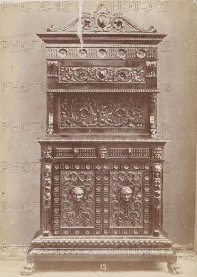 Decorated sideboard, No 13 Buffet, photograph, page, inscription: 'Ies. 160', inscription: 'Alto, 215. Largo, 111. Fondo, 49.', c. 1878 - c. 1885, photographer, paper, albumen print, height 131 mm x width 96 mm, Photo on unnumbered page, ornamental cupboard, A tall ornate wooden cabinet fills the frame, showing its upper decorative crest, carved panels, and base. The top features a curved carved pediment with floral and shell-like motifs and a central oval medallion, with a vertical lighter streak running down from near the crest. Below that is a horizontal frieze with circular rosette carvings and fluted details, followed by a recessed panel with intricately carved scrolling foliage and a central cartouche. The middle section has a projecting shelf with a row of small repeating rectangular dentils along its edge and a large carved panel beneath depicting swirling acanthus leaves and a stylized face at the center. Flanking columns with carved vertical ridges and small sculpted faces su...