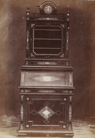 Ivory inlaid case, No 15 Mobile Avorio, photograph, page, inscription: 'Alto, 178. Largo, 79. Fondo, 55.', inscription: 'Ies. 360', c. 1878 - c. 1885, photographer, paper, albumen print, height 135 mm x width 97 mm, Photo on unnumbered page, ornamental cupboard, A tall ornate wooden cabinet photographed against a plain backdrop, showing upper and lower sections with decorative inlaid patterns and geometric borders; the top has a crowned pediment with a round medallion featuring a floral motif flanked by small urn finials, below which is a framed recessed display area with horizontal shelves and corner triangular panels filled with intricate scrollwork; a middle slanted panel displays a central ornamental cartouche surrounded by narrow borders, and the lower doors feature a large central diamond-shaped medallion with floral inlay and surrounding rectangular stepped panels; narrow vertical pilasters with repeating floral inlays run along the sides, and the cabinet rests on a short molded...