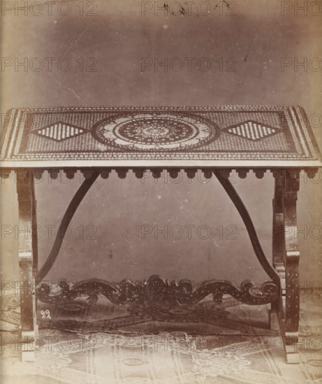 No 22 Tavola, Table with marquetry, photograph, page, inscription: 'Alta, 87. Larga, 122, Fonda 69.', inscription: 'Ies 200', c. 1878 - c. 1885, photographer, paper, albumen print, height 126 mm x width 109 mm, Photo on unnumbered page., table, A rectangular table with a patterned top featuring a central circular motif and two diamond-shaped checkerboard designs, supported by carved wooden legs and curved braces, with a decorative apron beneath the tabletop and a carved stretcher connecting the legs, all shown against a plain background and a rug partially visible on the floor.