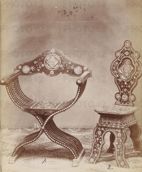 No 24 Poltrona, Sedia, Two types of chairs, photograph, page, inscription: 'No 1. Ies. 60., ' 2. 45', number: '1. 2.', inscription: 'Alta, 94. Larga, 70. Fonda, 36., ' 1m. '32. '35.', c. 1878 - c. 1885, photographer, paper, albumen print, height 127 mm x width 104 mm, Photo on unnumbered page., chair, A sepia-toned photograph showing two ornately decorated wooden chairs positioned on a patterned floor against a plain backdrop; the chair on the left has curved armrests, a decorative crest on the back with circular dot patterns, a curved slatted seat, and legs formed by interlacing curved slats with rows of round dot embellishments; the chair on the right features a tall, carved back with layered ornate shapes filled with dot motifs, a square seat with a patterned surface, and carved legs decorated with similar circular dot designs.