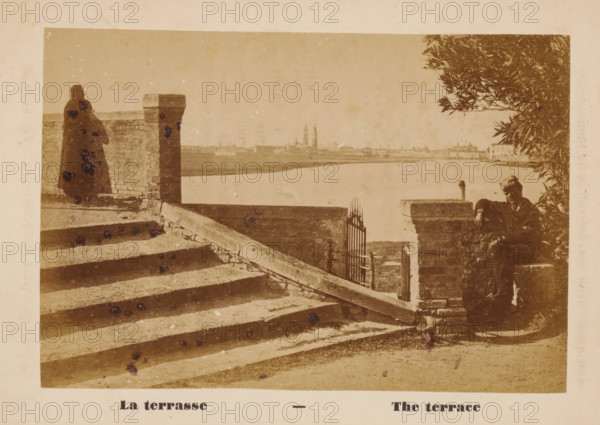 View of Venice from the quay of San Lazzaro degli Armeni, La terrasse, The terrace, photograph, page, San Lazzaro degli Armeni, c. 1874 - in or before 1879, photographer, paper, albumen print, height 88 mm x width 129 mm, Photo on unnumbered page opposite page 23., city-view in general, 'veduta', terrace, Venice, A sepia-toned scene shows wide stone steps leading down to a gated opening in a low stone wall with a diagonal stone beam across the steps; a tall stone pillar stands at the top of the steps beside a brick wall where a figure draped in a cloak stands facing the water; across the water a distant city skyline with spires and low buildings is visible under a pale sky; on the right a person in dark clothing sits on a stone ledge partially shaded by leafy branches of a tree, with the water and shoreline extending behind them.