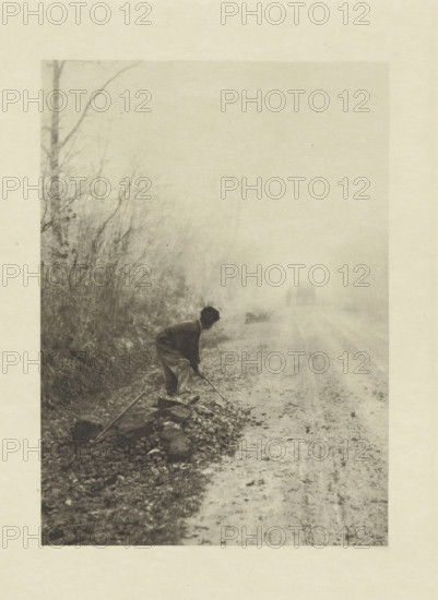 Worker along the road, Dans le brouillard, photomechanical print, photographer: Jean-Ferdinand Coste, cliché maker: Fillon & Heuse, printer (person): Charles Wittmann, Paris, c. 1891 - in or before 1896, printer (person), paper, photoaquatint, height 165 mm x width 118 mm, road, path, working class, laborers, A grainy monochrome scene shows a person bending forward near the edge of a rough road, holding a long tool and working among scattered stones and debris, with a line of bare trees and tangled underbrush along the roadside fading into a misty background and a faint silhouette of a horse-drawn vehicle visible down the road.