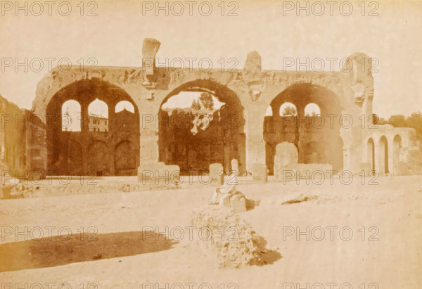 Ruin of the Temple of Peace in Rome, Roma, Foro Romano - tempio della pace, photograph, page, number: '1036.', Rome, c. 1883 - in or before 1888, photographer, paper, albumen print, height 100 mm x width 140 mm, Photo on page 137., landscape with ruins, Peace Temple, A faded photograph of a large stone structure with a series of tall rounded arches set on a raised platform, showing weathered surfaces and areas of erosion, with smaller archways visible within the larger arches and a few freestanding stone blocks and rubble scattered in front; a figure seated on a low stone in the foreground, indistinct in detail, and a patch of darker ground casting a shadow at the lower left, with a pale sky above.