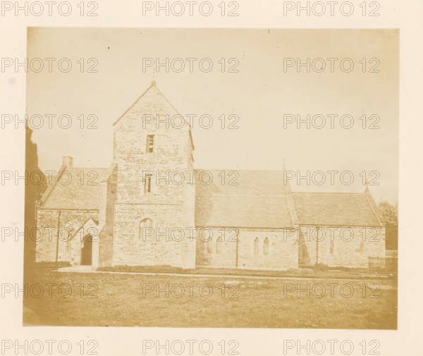 Exterior of a church, possibly in Canterbury, photograph, page, c. 1861 - in or before 1869, photographer, paper, albumen print, height 93 mm x width 117 mm, church (exterior), A sepia-toned photograph of a stone church building showing a central tower with a pitched roof and narrow windows, flanked by lower roofed sections with tiled sloping roofs and pointed arched windows, an arched doorway set into a projecting entrance on the left, stone walls with visible masonry texture, grassy foreground leading up to the building, and a plain sky occupying the upper portion of the image.