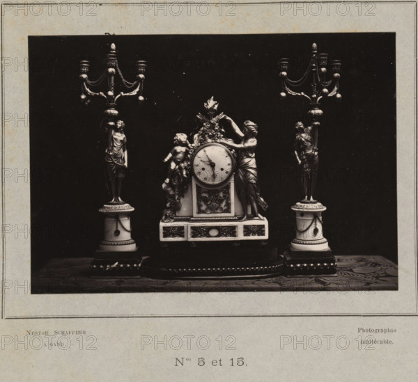 Clock and two candlesticks, photograph, page, inscription: 'Photographie, inaltérable.', number: 'PL. III., Nos 5 et 15.', photographer: Paul Joseph Nicolas Nestor Schaffers, 1873 - 1878, paper, carbon print, height 95 mm x width 134 mm, chandelier, candelabrum, clock, A black rectangular photograph mounted on a pale mat shows an ornate clock set flanked by matching candelabra on a patterned surface; the central clock features a round face with hands, sculptural floral and ribbon decorations, a reclining cherub figure on one side, and a draped female figure leaning against the clock on the other side, while each candelabrum rises from a columnar base topped with a standing draped female figure and branching arms with decorative chains and bulbous ornaments.