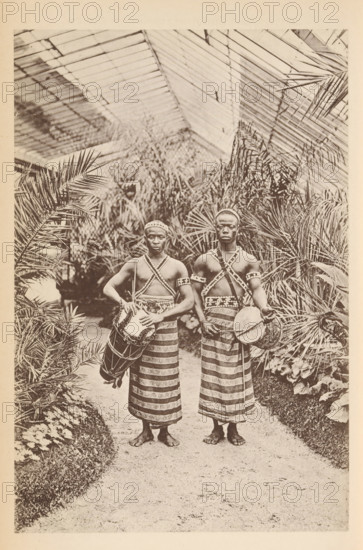 Portrait of two unknown men from Dahomey with drums in the Jardin d'acclimatation, Paris, Les Dahoméens au jardin d'acclimatation, page, photomechanical print, photographer: Maurice Bucquet, cliché maker: Rouillé, c. 1886 - in or before 1891, paper, collotype, photoprint, height 178 mm x width 119 mm, Print on unnumbered page., historical persons not known by name, drum (musical instrument), greenhouse, orangery, Jardin d'acclimatation, Two barefoot men stand side by side on a path lined with plants, each wearing patterned wrap skirts and crisscrossed beaded chest straps, headbands, and arm bands; both hold drums suspended by straps, with one drum held at the side and the other positioned in front, and a glass-roofed structure with large fronds and foliage fills the background.