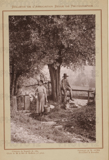 Un coin du Condroz (original title), Boy and a woman by a fence under a tree, Condroz, page, photomechanical print, inscription: 'Concours de Mr le Bon de Moffarts (Ir prix).', photographer: Armand baron de Moffarts, clichémaker: Wilhelm Otto, Brussels, c. 1879 - in or before 1884, printer (person), pasteboard, paper, collotype, photogravure, height 148 mm x width 98 mm, Print on unnumbered page., boy (child between toddler and youth), fence, wall, paling, farmers - AA - female farmer, farmer's wife, trees, A sepia-toned photograph mounted on a page shows a large tree with dense foliage dominating the upper portion and center. Beneath the tree, a young woman in a long skirt and blouse stands holding a metal pail at her side, looking slightly to her left. Nearby, a man wearing a jacket and wide-brimmed hat sits on a wooden rail fence with one leg bent and the other hanging down, facing forward. A dog lies on the ground near the man's feet. Grass, scattered stones, and small plants cover...