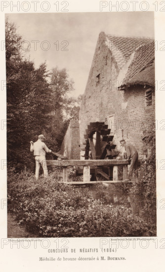 Two men adjust the water level at a watermill, in Eijsden, page, photomechanical print, inscription: 'Concours de négatifs (1884), Médaille de bronze décernée à M. Boumans.', photographer: Mathias Boumans, Eijsden, clichémaker: Goupil & Cie., Paris, c. 1879 - in or before 1884, printer (person), paper, Woodburytype, Woodburytypie, height 149 mm x width 98 mm, Print on unnumbered page., watermill, water mill, A sepia-toned vertical photograph shows an old brick building with a steep tiled roof and a thatched section, a wooden waterwheel attached to the building, and a wooden trough structure in front of the wheel. Two men stand by the trough facing the wheel; one man on the left wears light clothing and a cap and holds a long tool over his shoulder, the other man on the right leans forward wearing darker clothing and a hat. Dense foliage and trees occupy the left side and background, and grassy plants and low vegetation fill the foreground with a small reflective water surface near the ...
