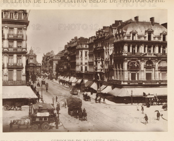 Street view in Brussels, page, photomechanical print, inscription: 'Concours de négatifs 1885', photographer: Henri Tournai, Brussels, cliché maker: workshop of Adolphe Braun & Cie., Dornach, c. 1880 - in or before 1885, printer (person), Mulhouse, paper, Woodburytype, height 118 mm x width 157 mm, Print on unnumbered page., street, A wide street scene shows multiple ornate multi story buildings with decorative façades and balconies lining both sides, a corner building with a rounded roof and sculpted window surrounds visible on the right, rows of awnings extending over shopfronts beneath the upper floors, and a distant churchlike building with a pediment visible along the left side. The street is populated with horse-drawn carriages and wagons, drivers and pedestrians walking or standing, a large load of bundled material on a cart moving down the road, several street lamps along the pavement, and small kiosk or booth near the foreground curb. The image is monochrome with a slightly fa...