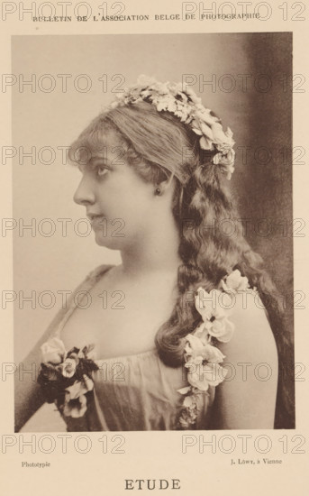 Etude, Portrait of an unknown woman with a flower wreath, page, photomechanical print, photographer: Josef Löwy, Vienna, c. 1883 - in or before 1888, printer (person), Lower Austria, paper, collotype, photoprint, height 148 mm x width 104 mm, Print on unnumbered page., flowers, adult woman, historical persons not known by name - BB - woman, A sepia-toned portrait of a young woman shown in profile facing left, with long wavy hair cascading over her shoulder and adorned with a garland of flowers on her head and along her dress strap; she wears a sleeveless, low-cut gown with floral decorations at the bodice, a small earring, and a calm, neutral expression against a plain, softly shaded background.
