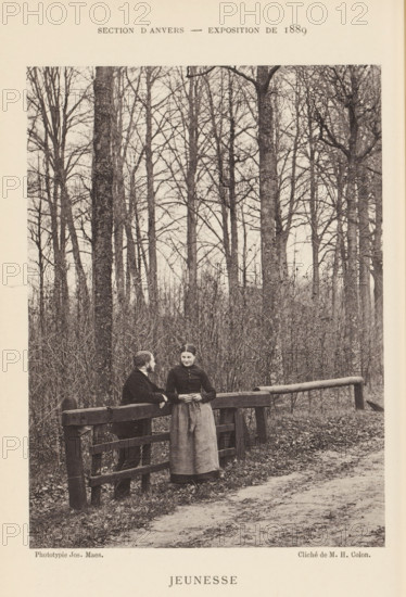Jeunesse, Man and woman, talking in a forest, page, photomechanical print, inscription: 'Section d'Anvers - Exposition de 1889', photographer: Henri Colon, cliché maker: Joseph Maes, printer (person): Joseph Maes, Antwerp, c. 1883 - in or before 1888, Henri Colon, Joseph Maes, photographer, cliché maker, printer (person), paper, collotype, photoprint, height 156 mm x width 115 mm, Print on unnumbered page., forest path or lane, lovers, courting, flirting, A black and white photograph shows a wooded scene with tall leafless trees and dense underbrush. In the foreground there is a wooden fence running parallel to a dirt path, with a horizontal log barrier extending from the fence into the woods. A man and a woman stand beside the fence; the man leans on the top rail facing the woman, and the woman stands upright with her hands clasped in front of her. The man wears a dark jacket and trousers and has short hair and facial hair, while the woman wears a dark bodice with a long skirt and her...