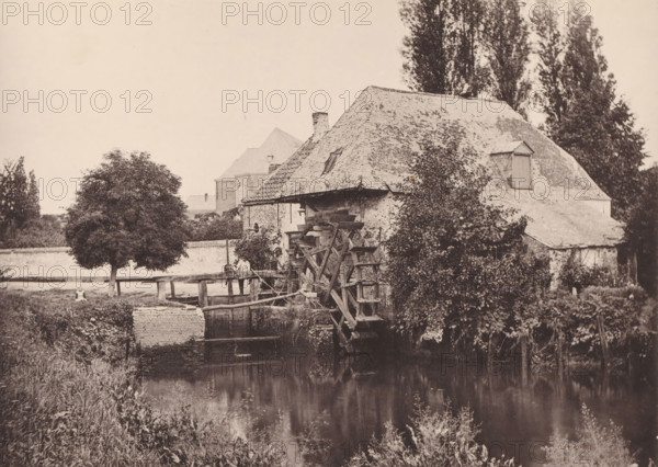 Moulin and Campine, Watermill in Kempenland, page, photomechanical print, inscription: 'Concours de négatifs, 1888, Médaille de Vermeil', photographer: Henri Schleusner, Kempen (Belgium), cliché maker: Joseph Maes, Antwerp, c. 1883 - in or before 1888, printer (person), paper, collotype, photoprint, height 110 mm x width 154 mm, Print on unnumbered page., watermill, A sepia-toned photograph showing an old stone building with a steep tiled roof and a small dormer, partially covered by climbing vegetation, beside a wooden waterwheel structure connected to a low stone wall and a narrow channel of water; tall trees rise behind the building and a rounded tree stands in a grassy area beside a wooden fence, with overgrown grasses and plants along the water’s edge and a reflection visible in the water.