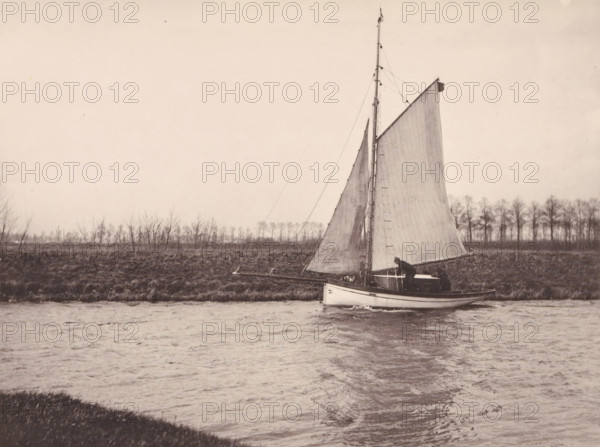 Sailing ship on the Leie, page, photomechanical print, inscription: 'Concours de négatifs, 1888, Médaille de Bronze', photographer: Guillaume van Assche, Flanders, cliché maker: Joseph Maes, Antwerp, 1887-04-24, printer (person), paper, collotype, photoprint, height 120 mm x width 159 mm, Print on unnumbered page., sailing-ship, sailing-boat, Leie, A sepia-toned photograph showing a sailboat on a body of water close to a grassy bank, with the boat’s tall mast and large sail raised and rigging lines visible, a person standing near the stern tending the vessel, a shoreline with low vegetation and a row of bare trees in the background, and ripples and reflections on the water surface in the foreground.