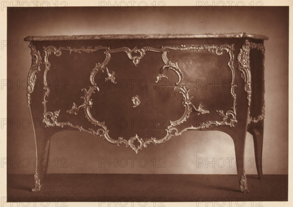 Chest of drawers in the style of Louis XV, from the collection of Christopher Beckett Denison, page, photomechanical print, number: '252', c. 1880 - in or before 1885, paper, Woodburytype, height 123 mm x width 178 mm, ornamental cupboard (wood and other phytogenic material), A sepia-toned photograph of an ornate wooden table with a polished top and elaborately carved decorative motifs along its front and legs, featuring flowing scrollwork, floral elements, and curved framing around a central panel, the table stands on slender cabriole legs with carved knees and feet, and the surface shows subtle reflections and a slightly darker backdrop behind the furniture.