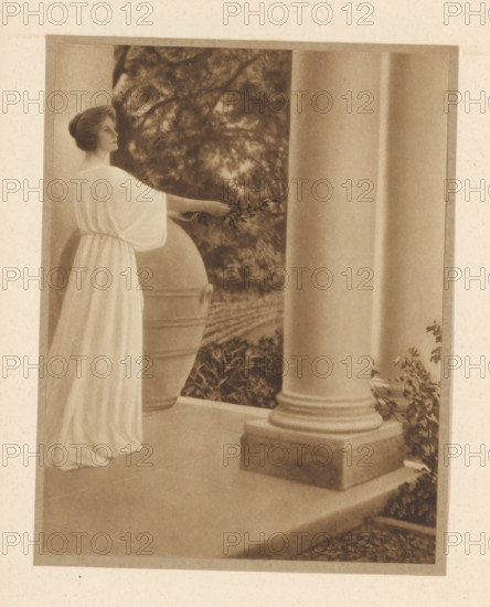Woman with laurel branch at a temple, page, photomechanical print, copyright notice: 'C', maker: Adelaide Hanscom Leeson, c. 1906 - in or before c. 1916, paper, photoaquatint, height 163 mm x width 125 mm, Print on unnumbered page. Adelaide Hanscom Leeson manipulated her glass negatives to create a painterly effect., adult woman, trees: laurel (branch, stick), A sepia-toned photograph showing a woman in a flowing light-colored gown standing on a porch beside a large classical column and a rounded decorative urn; she is in profile with her hair styled close to her head and her arm extended holding a small leafy branch toward the space between the urn and the column, while behind her a backdrop of dense foliage and trees is visible and the porch floor and column base occupy the foreground.