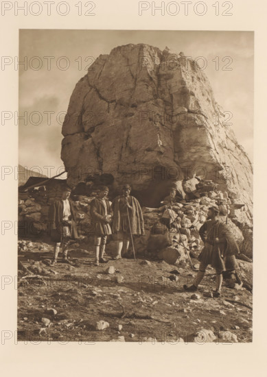 Les bergers de Parnasse et leurs hôtes, Shepherds on Parnassus, page, photomechanical print, photographer: Frédéric Boissonnas, Parnassus, cliché maker: Meisenbach, Riffarth & Cie., Berlin, c. 1905 - in or before 1910, paper, photoaquatint, height 290 mm x width 224 mm, Photo on page 96., herding, herdsman, herdswoman, shepherd, shepherdess, cowherd, A large rocky outcrop fills the center and upper portion of the image, with rough, cracked surfaces and light and shadow defining its contours. At the base of the rock is a low stone wall or pile of stones forming a rough shelter area. Five people wearing heavy cloaks and traditional-looking garments stand and sit near the stone wall; some hold long staffs or walking sticks and one appears seated among the stones. The ground in the foreground is uneven and strewn with rocks and small boulders. The overall image has a monochrome, sepia-toned appearance.