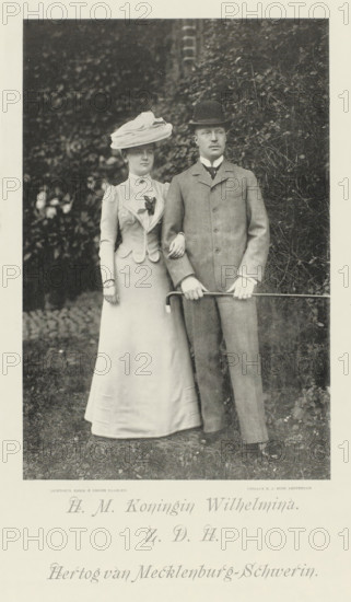 Portrait of Wilhelmina, Queen of the Netherlands, and Hendrik of Mecklenburg-Schwerin, photomechanical print, Portrait of Wilhelmina and Hendrik. Their names and titles in the lower margin., cliché maker: Emrik & Binger, Haarlem, publisher: Nicolaas Jan Boon, Amsterdam, 1900, photographer, paper, collotype, photogravure, height 346 mm x width 229 mm, The title mentioned here refers to the highest title obtained by the sitter, not necessarily to the title the sitter bears in this image., Person, Hendrik (Prince of the Netherlands (1876-1934)), Wilhelmina of the Netherlands, A black-and-white photograph of a woman and a man standing outdoors against a background of dense foliage, the woman on the left wearing a long light-colored dress with a fitted jacket, a large wide-brimmed hat with layered fabric, a decorative floral detail on her chest, and her arm linked through the man's arm; the man on the right wearing a three-piece suit with a jacket buttoned at the front, a collared shirt wit...