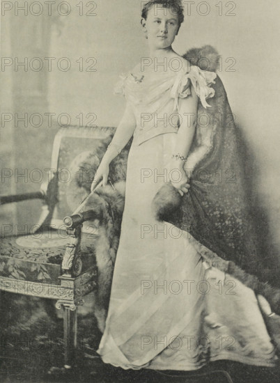 Portrait of Wilhelmina, Queen of the Netherlands, photomechanical print, Portrait of Wilhelmina. She stands next to a chair. In the lower margin her name and title., photographer: Heinrich Wilhelm Wollrabe, clichémaker: anonymous, Netherlands, 1897, paper, collotype, photoprint, height 347 mm x width 217 mm, The title mentioned here refers to the highest title obtained by the sitter, not necessarily to the title the sitter bears in this image., Person, Wilhelmina of the Netherlands, A sepia-toned portrait of a woman standing beside an ornately carved upholstered chair, wearing a floor-length gown with an off-the-shoulder bodice and delicate draped fabric at the sleeves, a fur-trimmed cloak draped over one shoulder and trailing toward the floor, bracelets on both wrists, one hand resting on the chair arm and the other holding the fur, her hair styled up, and the chair featuring decorative carved details and patterned upholstery.