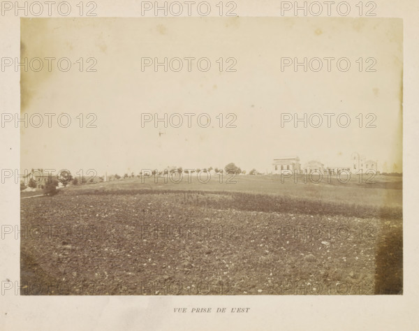 View of the observatory in Besançon, Vue prize de l'est, photograph, page, photographer: M. Lebeuf, Besançon, c. 1882 - in or before 1892, M. Lebeuf, photographer, paper, albumen print, height 160 mm x width 221 mm, observatory (, astronomy), meteorological observatory, Observatoire de Besançon, A wide sepia photograph showing a plowed field in the foreground with visible soil clumps and scattered stones, a gently sloping strip of grass and sparse vegetation above the field, and a low horizon line dotted with trees and small buildings including several houses and larger classical-style structures with columns and pitched roofs; the sky occupies much of the upper area and appears pale and empty with a few faint spots and blemishes on the photographic surface.