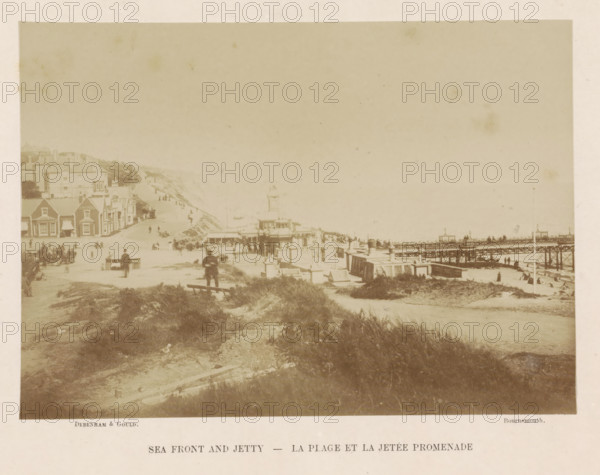 Sea front and jetty, photograph, page, photographer: Debenham & Gould, Bournemouth, c. 1876 - in or before 1886, paper, albumen print, height 144 mm x width 202 mm, coast, quay, A faded sepia photograph showing a coastal scene with a sandy foreground and tufts of grass leading toward a promenade and buildings along the shoreline; there are houses with pitched roofs and awnings on the left, a central pavilion or towered structure near the water, a wooden jetty extending out from the right with people visible along it and on the beach, scattered groups of people and horse-drawn vehicles along the promenade, and a gently sloping hillside with additional buildings receding into the distance under a pale sky.