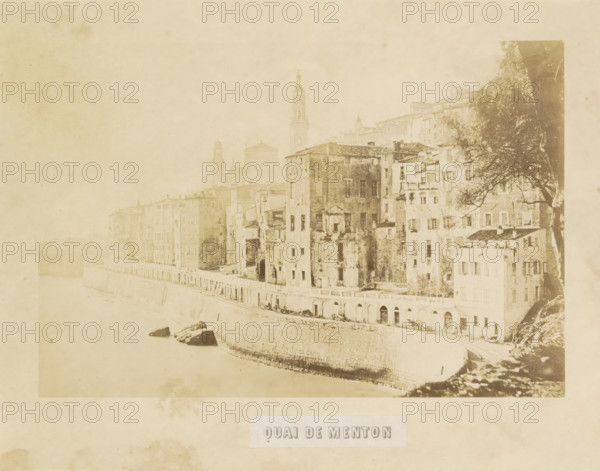 Quai de Menton, View of the quay of Menton, photograph, page, photographer: Louis-Alphonse Davanne, Menton, c. 1852 - in or before 1862, Provence-Alpes-Côte d'Azur, paper, albumen print, height 122 mm x width 182 mm, façade (of house or building), quay, A faded sepia photograph shows a riverside view with a stone embankment curving along the water and tall, weathered buildings rising directly from the embankment, their facades marked by windows, balconies, and patches of wear; a tower and church silhouettes are visible behind the row of buildings, and a large tree with spreading branches appears on the right side near the structures, while rocks rest near the water's edge and the overall image has an aged, grainy texture.