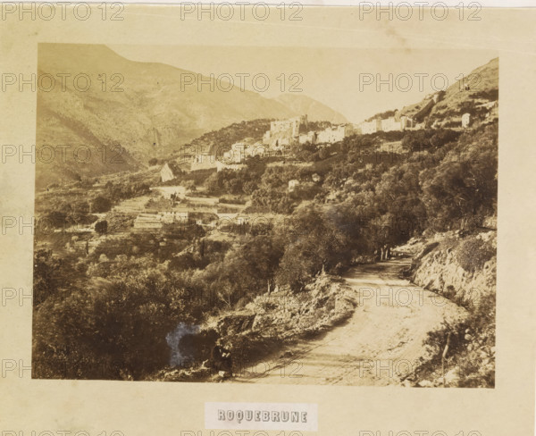 View of Roquebrune-Cap-Martin, Roquebrune, photograph, page, photographer: Louis-Alphonse Davanne, Roquebrune-Cap-Martin, c. 1852 - in or before 1862, Provence-Alpes-Côte d'Azur, paper, albumen print, height 127 mm x width 177 mm, Photo for page 36., city-view in general, 'veduta', A sepia-toned landscape photograph showing a winding dirt road in the foreground bordered by rocky embankments and scattered trees, with a person seated at the road's edge near a shaded area. The middle ground contains terraced fields, stone buildings, and clusters of trees rising toward a hillside village with closely packed houses and a taller structure that stands above the others. The background features layered mountain slopes fading into the distance under a pale sky, and the entire image is mounted on a lighter paper border.