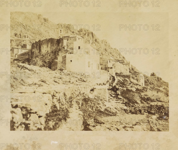 Ste. Agnes, View of Sainte-Agnès, photograph, page, photographer: Louis-Alphonse Davanne, Sainte-Agnès, c. 1852 - in or before 1862, Provence-Alpes-Côte d'Azur, paper, albumen print, height 137 mm x width 198 mm, Photo for page 40., city-view in general, 'veduta', Sainte-Agnes, A faded, sepia-toned photograph of a rocky hillside with a cluster of stone buildings and ruins built into the slope, showing rough stone walls, stairways, and terraced paths leading upward; one taller building with a small squared tower and simple rectangular openings is visible among lower structures, and sparse leafless trees and scattered rocks are present on the uneven terrain beneath a pale, textured sky.