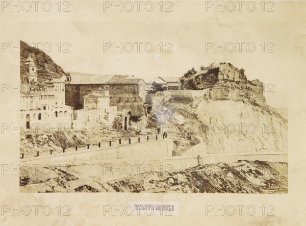 View of Ventimiglia, Vintimille, photograph, page, photographer: Louis-Alphonse Davanne, Italy, c. 1852 - in or before 1862, paper, albumen print, height 125 mm x width 197 mm, city-view in general, 'veduta', Ventimiglia, A sepia-toned photograph shows a cliffside town with stone buildings perched along steep rocky terrain, including a multi-story structure with arched openings and a taller tower with a domed roof on the left, a long stone wall running horizontally across the lower part of the scene with evenly spaced short posts along its top, rough rocky slopes and terraces beneath the wall, and large layered rock formations and vegetation on the right where the cliff face drops away.