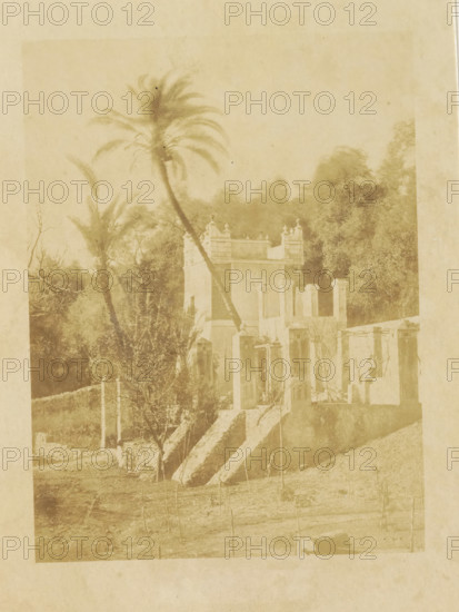 Jardins Moreno Bordighera, Part of the Moreno Gardens in Bordighera, photograph, page, photographer: Louis-Alphonse Davanne, Bordighera, c. 1852 - in or before 1862, paper, albumen print, height 176 mm x width 132 mm, Photo for page 51., garden, Ventimiglia, A faded sepia photograph showing a small building with ornate parapets and pillars, a set of worn stone steps leading up to doorways, and partial ruined walls; a tall, leaning palm tree rises beside the building with fronds splaying at the top, and additional trees and dense foliage fill the background; the foreground shows a rough grassy area with sparse young stakes or saplings and a low stone wall segment, all rendered in soft, aged tones.