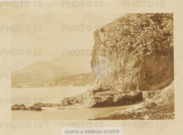 Grotte & rochers St. Louis, Rocks along the coast of France, photograph, page, photographer: Louis-Alphonse Davanne, France, c. 1852 - in or before 1862, paper, albumen print, height 129 mm x width 194 mm, Photo for page 51., rocky coast, A sepia-toned photograph showing a large rocky cliff dominating the right side, with textured rock faces, crevices, and vegetation patches; below the cliff are layered rocky outcrops and a shoreline with scattered stones leading into calm water; in the distance on the left a hazy town sits along the water with building rooftops and a church steeple visible against low rolling hills; the sky and distant hills appear pale and faded, and the image has an overall aged, mottled paper background.