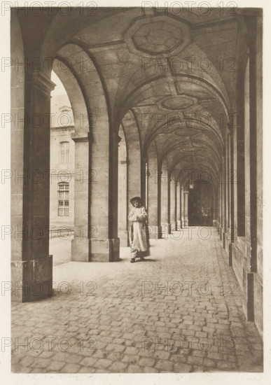 Monk in an arcade, page, photomechanical print, photographer: Henri Magron, France, cliché maker: Paul Dujardin, c. 1885 - in or before 1896, paper, photoaquatint, height 206 mm x width 140 mm, monk(s), friar(s), arcade, architecture, A long covered stone arcade with a vaulted ceiling and repeating arches receding into the distance, stone columns on one side and a wall with window openings on the other, a cobblestone floor, and a person standing near one of the central columns wearing a long coat and a hat while holding something in front of their chest.