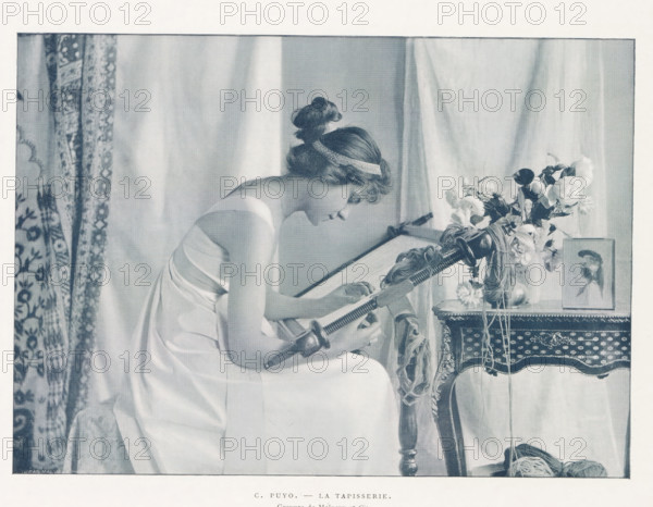 Woman behind a loom, La tapisserie, page, photomechanical print, name: 'Jean Malvaux PH', photographer: Constant Puyo, cliché maker: Malocuy et Cie, cliché maker: Malvaux et Cie, c. 1889 - in or before 1899, paper, autotypie, height 150 mm x width 210 mm, weaving-loom, A seated woman in a light sleeveless dress leans forward, her hair wrapped with a ribbon and gathered in a loose bun, as she works with a long handled tool over a frame; beside her an ornate table holds a vase of mixed flowers and a framed portrait, and yarn and cords hang and lie near the table; patterned drapery and plain curtains form the background.