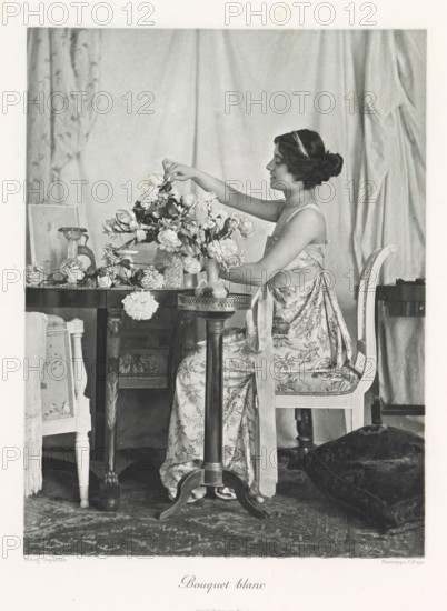 Flower arranging woman in an interior, Bouquet blanc, page, photomechanical print, photographer: Constant Puyo, France, cliché maker: Paul Dujardin, printer (person): Charles Wittmann, Paris, c. 1886 - in or before 1896, printer (person), paper, photoaquatint, height 218 mm x width 165 mm, adult woman, arranging flowers, interior, representation of a building, A seated woman in a patterned dress with a ribbon tied around her waist arranges a large bouquet of flowers on a wooden table, her hair pinned up with a headband and her profile visible as she reaches toward the blooms; the table holds various vases and scattered blossoms, and an ornate chair with a cushioned seat supports her while a small round pedestal table stands in front; draped fabric hangs in the background and a patterned rug covers the floor, with a dark cushion or piece of fabric resting nearby.