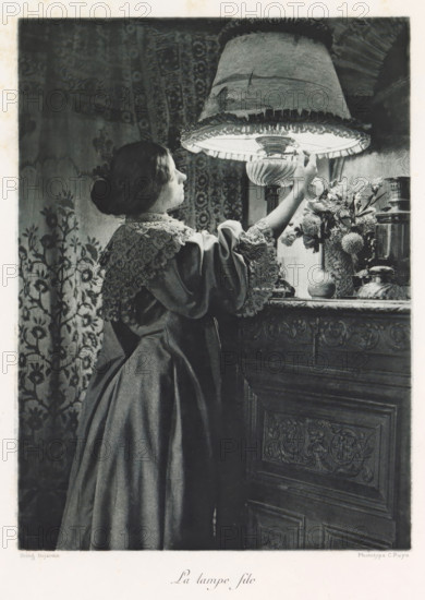 Woman turns on a lamp, La lampe file, page, photomechanical print, photographer: Constant Puyo, France, cliché maker: Paul Dujardin, printer (person): Charles Wittmann, Paris, c. 1886 - in or before 1896, printer (person), paper, photoaquatint, height 218 mm x width 160 mm, adult woman, lighting, lamps, A woman in a long dark dress with a lace collar and sleeves stands in profile reaching up to adjust a large fringed lampshade; her hair is pulled back in a bun and she faces the illuminated interior of the lamp. The lamp has a decorative base and sits on a carved wooden surface beside a vase of flowers and other small objects. Behind her is a patterned textile or wallpaper with floral motifs. The scene is rendered in grayscale with soft lighting highlighting the woman's face and the lamp area.