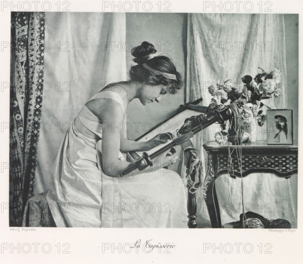 La tapisserie, Weaving woman in an interior, page, photomechanical print, photographer: Constant Puyo, France, cliché maker: Paul Dujardin, printer (person): Charles Wittmann, Paris, c. 1886 - in or before 1896, printer (person), paper, photoaquatint, height 147 mm x width 197 mm, adult woman, weaving-loom, interior, representation of a building, weaving, A seated woman in a light-colored draped dress leans forward, her hair styled in a bun with a decorative band, as she works with a wooden tool held across her lap; to her right is an ornate table partly covered by fabric, with a vase of assorted flowers and a framed portrait propped on the table, and skeins of yarn hanging from the tool and resting near the table leg; patterned curtains and plain drapery form the background.