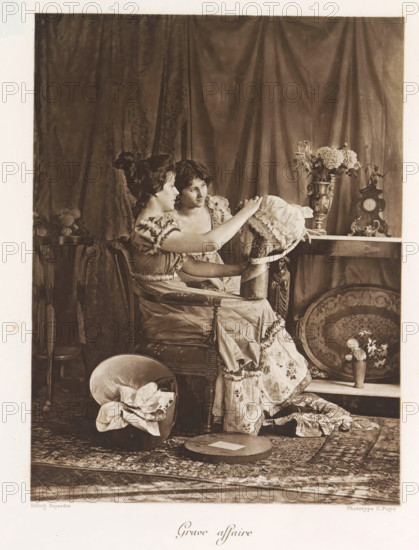 Grave affair, Two women look at a hat in an interior, page, photomechanical print, photographer: Constant Puyo, France, cliché maker: Paul Dujardin, printer (person): Charles Wittmann, Paris, c. 1886 - in or before 1896, printer (person), paper, photoaquatint, height 202 mm x width 150 mm, adult woman, fan, interior, representation of a building, Two young women sit closely on an upholstered chair in front of a draped curtain, one woman facing slightly sideways and the other leaning toward her while both focus on a bonnet that the nearer woman holds; the women wear patterned dresses with ruffled sleeves and layered skirts. Around them are decorative objects including a table with a tall ornate vase of flowers and an antique clock, a round decorative tray propped against the shelf, and a small pedestal with a figurine. On the floor in front of them a hat box contains folded fabric and a flat round hat box lid rests nearby on a patterned rug. The scene is sepia-toned with soft lighting a...