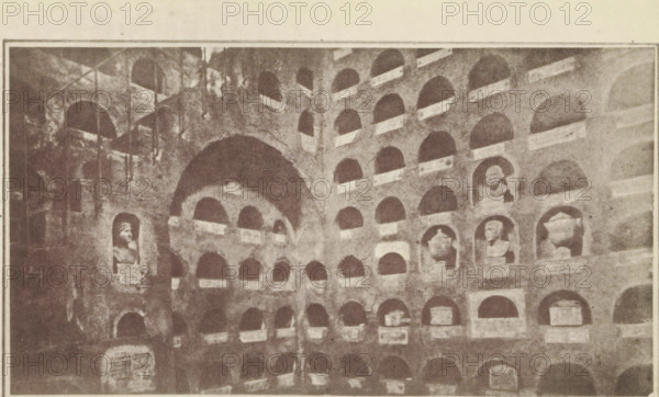 Interior of the columbarium of the Vigna Codini, Columbaria Tomb of 'The officers of Caesar's household', page, photomechanical print, number: 'Plate II', c. 1867 - in or before 1877, paper, photoaquatint, height 220 mm x width 140 mm, niche-tomb, wall-tomb, Rome, Columbarium of the Vigna Codini, A sepia-toned photograph of a wall filled with rows of recessed niches, many of which contain sculpted busts and urns, with several niches left empty; the wall shows arches and a large central archway, rough stone texture, and varying light and shadow across the surfaces, and a few vertical elements like poles or supports visible on the left side.