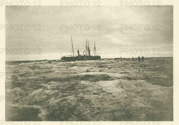 The Louise and the Varna moored in a patch of ice. Aug 17 1882, The Dutch Polar Expedition 1882-1883 published by the care of the Lieutenant Commander 1st class BJG Volck, photographer, photographer: H. Ekama, writer (clerical worker): Maurits Snellen, printer (person): E. Diener, London, publisher: Bosch & Zoon, LE, Balthasar Jean George Volck, c. 1886, writer (clerical worker), printer (person), paper, collotype, light printing on photo paper on cardboard, height 0 mm x width 0 mm, A grainy monochrome scene showing a shoreline with rough, textured rock surfaces in the foreground leading toward a distant horizon, a large ship with tall masts and a central smokestack positioned near the horizon line, several small human silhouettes standing on the rocks near the ship, a flat sky with subtle cloud banding above, and shallow pools of water among the rocky foreground.