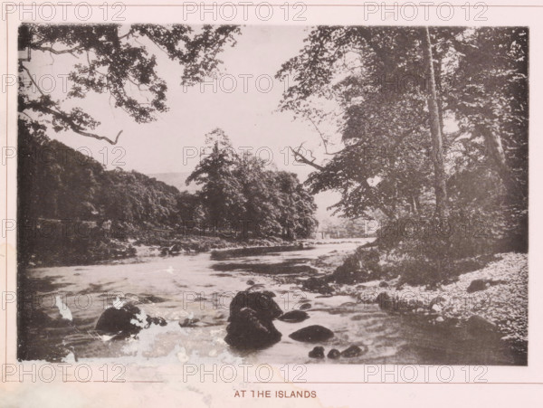 ... at the islands, page, photomechanical print, c. 1890 - c. 1900, textile materials, linen (material), collotype, photoprint, height 102 mm x width 152 mm, island, river, A sepia-toned landscape photograph showing a river flowing toward the foreground with visible ripples and scattered rocks emerging from the water, tree-lined banks on both sides with dense foliage and tall trunks, an overhanging canopy of branches and leaves framing the upper edges of the image, a pebbled or rocky shoreline on the right, and distant trees and hills partially visible through a light sky.