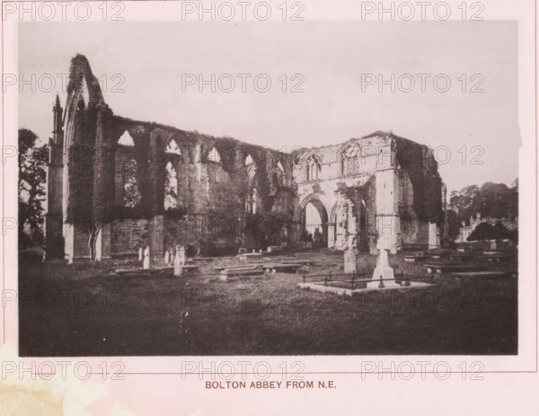 Bolton Abbey from NE, page, photomechanical print, c. 1890 - c. 1900, textile materials, linen (material), collotype, photoprint, height 102 mm x width 152 mm, ruin of church, monastery, Yorkshire, Wharfedale, Bolton Abbey, A sepia-toned photograph shows the ruins of a stone abbey with tall arched openings and partially collapsed walls surrounded by a grassy burial ground with upright and flat grave markers and low tombs; trees appear behind and beside the ruins and vegetation climbs parts of the remaining structure, while the sky above is pale and empty.