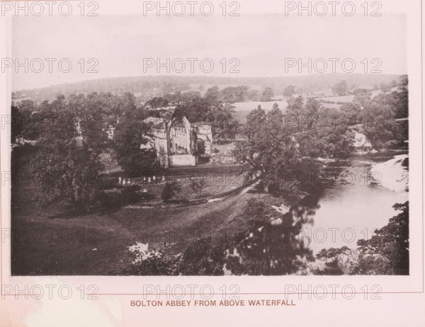 Bolton Abbey from above waterfall, page, photomechanical print, c. 1890 - c. 1900, textile materials, linen (material), collotype, photoprint, height 102 mm x width 152 mm, ruin of church, monastery, Yorkshire, Wharfedale, Bolton Abbey, A wide landscape photograph showing a river running along the right side with visible water flow and a lighter riverbank area, dense trees and foliage along both banks, open grassy land in the foreground, a large ruined stone building with a tall arched window and partial walls set among trees near the river, scattered small bright figures on the grass near the ruins, rolling wooded hills in the background, and a pale sky above.