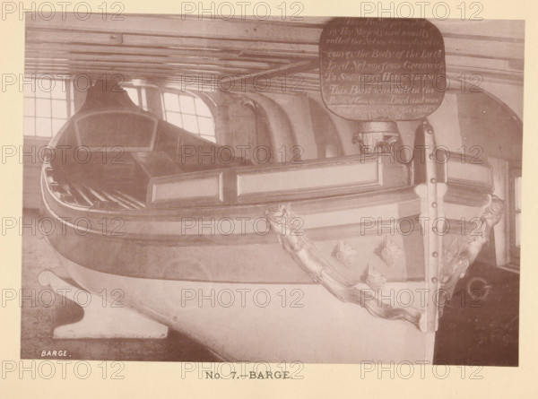 Barge, page, photomechanical print, number: 'No. 7.', c. 1881 - in or before 1891-05, paper, collotype, photoprint, height 112 mm x width 160 mm, boat propelled by man or animal, parts of ship's interior, A sepia-toned photograph of a wooden barge interior showing a carved bow with decorative scrollwork and shell motifs along its side, a polished rail with rectangular paneling, an elevated prow area with a curved inner seating or floor structure, a tall vertical post topped by a rounded plaque, a low beamed ceiling overhead, and mullioned windows letting in light in the background.