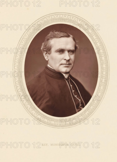 The rev. monsignor Capel, page, photomechanical print, photographer: Lock & Whitfield, cliché maker: anonymous, c. 1871 - in or before 1876, paper, Woodburytype, Woodbury Process, height 115 mm x width 89 mm, Person, Thomas John Capel, priest (Roman Catholic), historical persons (portraits and scenes from the life) ((full) bust portrait), An oval portrait of a middle-aged man with short wavy hair and a composed expression set against a plain dark background, the man wearing a dark clerical garment with a high collar and decorative piping, framed by an ornate oval border with intricate repeating leaf and scroll motifs on a light textured paper.