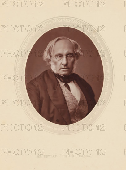 Sir Edward Shepherd Creasy, page, photomechanical print, photographer: Lock & Whitfield, cliché maker: anonymous, c. 1871 - in or before 1876, paper, Woodburytype, height 115 mm x width 89 mm, Person, Edward Creasy, historical persons (portraits and scenes from the life) ((full) bust portrait), An oval sepia portrait of an older man with sideburns and round eyeglasses set against a darker oval background, framed by an ornate light-colored decorative border, the man wearing a dark jacket over a patterned waistcoat and a white shirt with a dark cravat, his hair parted and swept to the sides, his expression neutral, the portrait set on a light textured sheet.