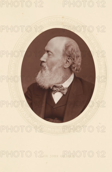 Sir John Gilbert, page, photomechanical print, photographer: Lock & Whitfield, cliché maker: anonymous, c. 1871 - in or before 1876, paper, Woodburytype, Woodbury Process, height 115 mm x width 89 mm, Person, John Gilbert, portrait, self-portrait of painter, historical persons (portraits and scenes from the life) ((full) bust portrait), A sepia-toned oval portrait of an older man shown in profile facing left, with a full white beard and mustache, receding hairline and curly hair at the back of his head, wearing a dark suit jacket and waistcoat over a white shirt and a patterned bow tie, set against a darker oval background and surrounded by a decorative pale border on a light sheet.