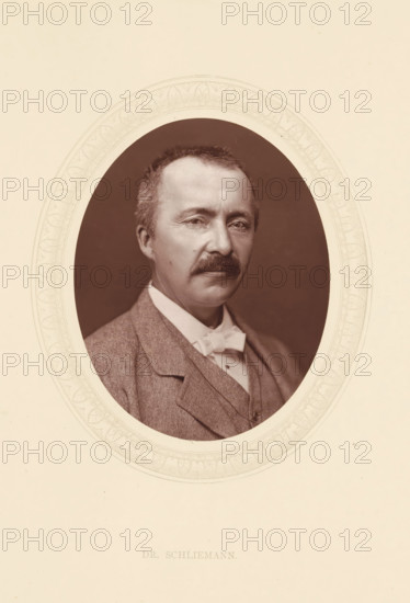 Dr. Schliemann, page, photomechanical print, photographer: Lock & Whitfield, cliché maker: anonymous, c. 1871 - in or before 1876, paper, Woodburytype, Woodbury Process, height 115 mm x width 89 mm, Person, Heinrich Schliemann, explorer, historical persons (portraits and scenes from the life) ((full) bust portrait), A sepia-toned oval portrait of a middle-aged man with short hair and a mustache, shown from the chest up and wearing a suit jacket, waistcoat, high-collared shirt, and a bow tie, set against a dark oval background and surrounded by a decorative light-colored oval frame on a pale rectangular sheet.
