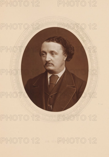 Dr. Stainer, page, photomechanical print, photographer: Lock & Whitfield, cliché maker: anonymous, c. 1873 - in or before 1878, paper, Woodburytype, Woodbury Process, height 115 mm x width 89 mm, Person, John Stainer, portrait of composer, historical persons (portraits and scenes from the life) ((full) bust portrait), A sepia portrait of a man shown from the chest up within an oval frame, wearing a dark suit jacket with wide lapels, a waistcoat, a white shirt, and a tie; he has short curly hair swept back, a mustache, and a contemplative facial expression, with a plain dark background inside the oval and a decorative patterned border around the oval set on a light beige background.